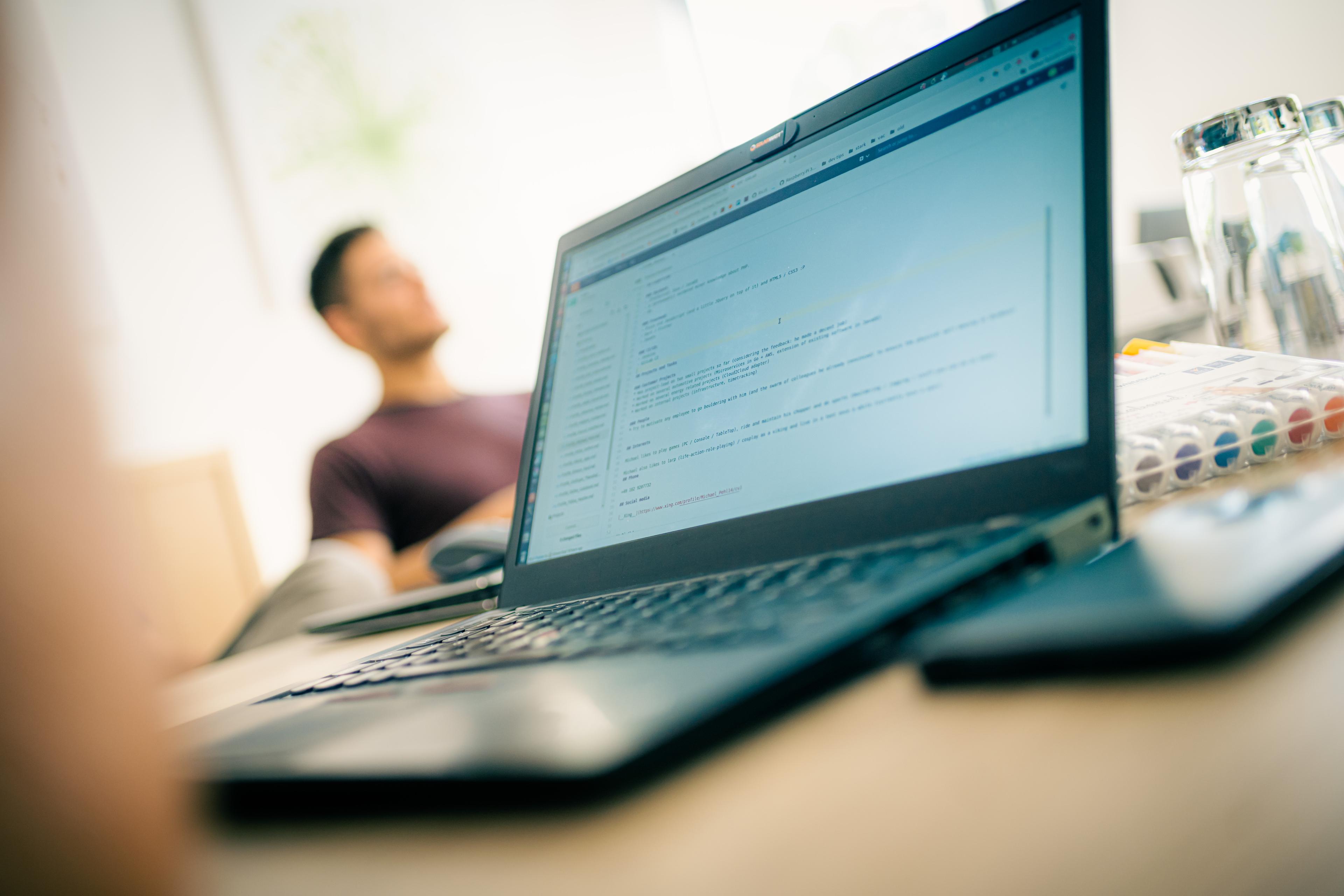 Bild von einem Notebook aktiver IDE, Sourcecode und einer verschwommenen Person im Hintergrund